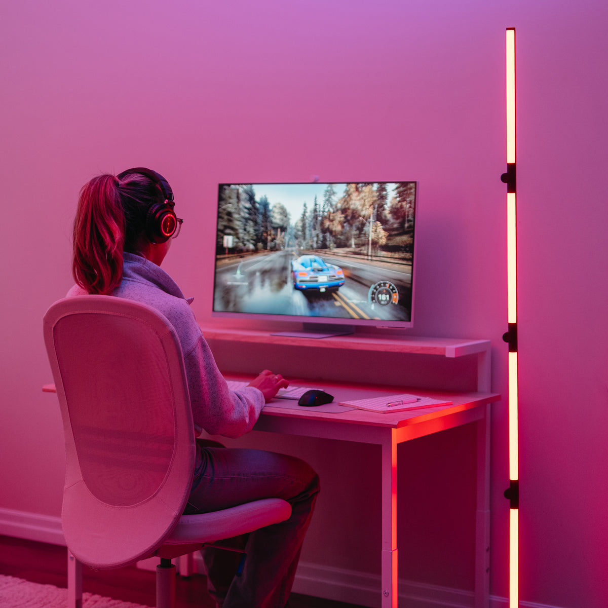 A person wearing headphones sits at a desk playing a racing video game on a monitor. The room is lit with pink and red LED lights, creating a colorful, modern gaming setup.