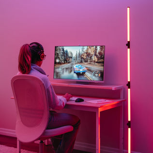 A person wearing headphones plays a racing game at a desk, illuminated by the Artika Fitch smart foldable RGB LED gaming floor lamp in a modern, minimalist room.
