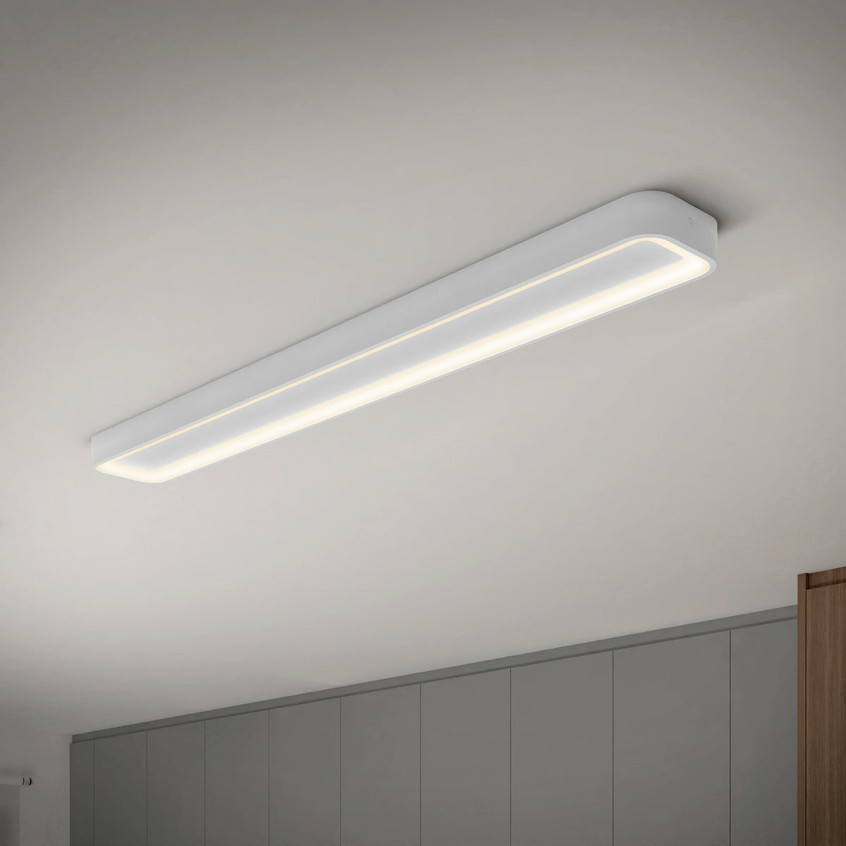 A modern, rectangular LED ceiling light fixture is switched on, emitting a soft, even glow in a minimalist room with light gray walls and a white ceiling.