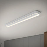 A modern, rectangular LED ceiling light fixture is switched on, emitting a soft, even glow in a minimalist room with light gray walls and a white ceiling.