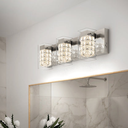 A modern bathroom wall light fixture with three cube-shaped crystal lights on a metallic base, mounted above a large mirror. The bathroom has white marble walls and a rainfall showerhead.