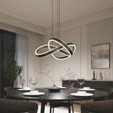 A modern dining room with a round black table set for four, featuring white plates, wine glasses, and a minimalist centerpiece. An abstract, intertwined LED chandelier hangs above. Contemporary decor and wall art complete the scene.