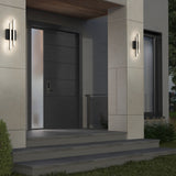A modern house entrance with a dark gray front door, frosted glass side panel, concrete steps, wall-mounted lights, and a large window to the right. The entryway is surrounded by beige walls and greenery.