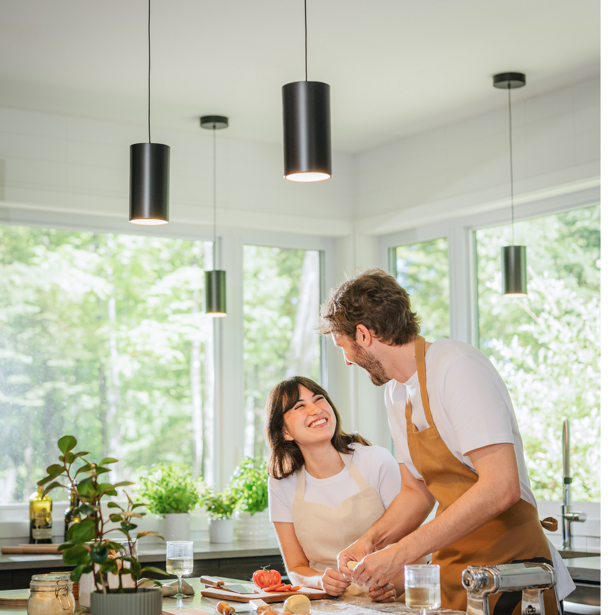 Two people cook beneath an Artika Alton mini LED pendant in a bright, modern kitchen with big windows, both smiling in aprons.