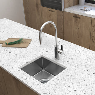 A modern kitchen countertop features the Artika Plaza 15 in. single bowl stainless steel bar sink with a curved faucet. A wooden board with chopped green onions sits beside the sink, and wood cabinets are visible in the background.