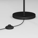 A close-up of a black floor lamp base with a cord extending out and connecting to a round, black foot switch on a white surface.
