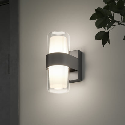 The Artika Colton modern LED indoor-outdoor wall light 5 CCT, featuring a cylindrical frosted glass shade with a black metal band, is mounted on a light wall and casts a soft glow. A plant appears partially in the upper right corner.