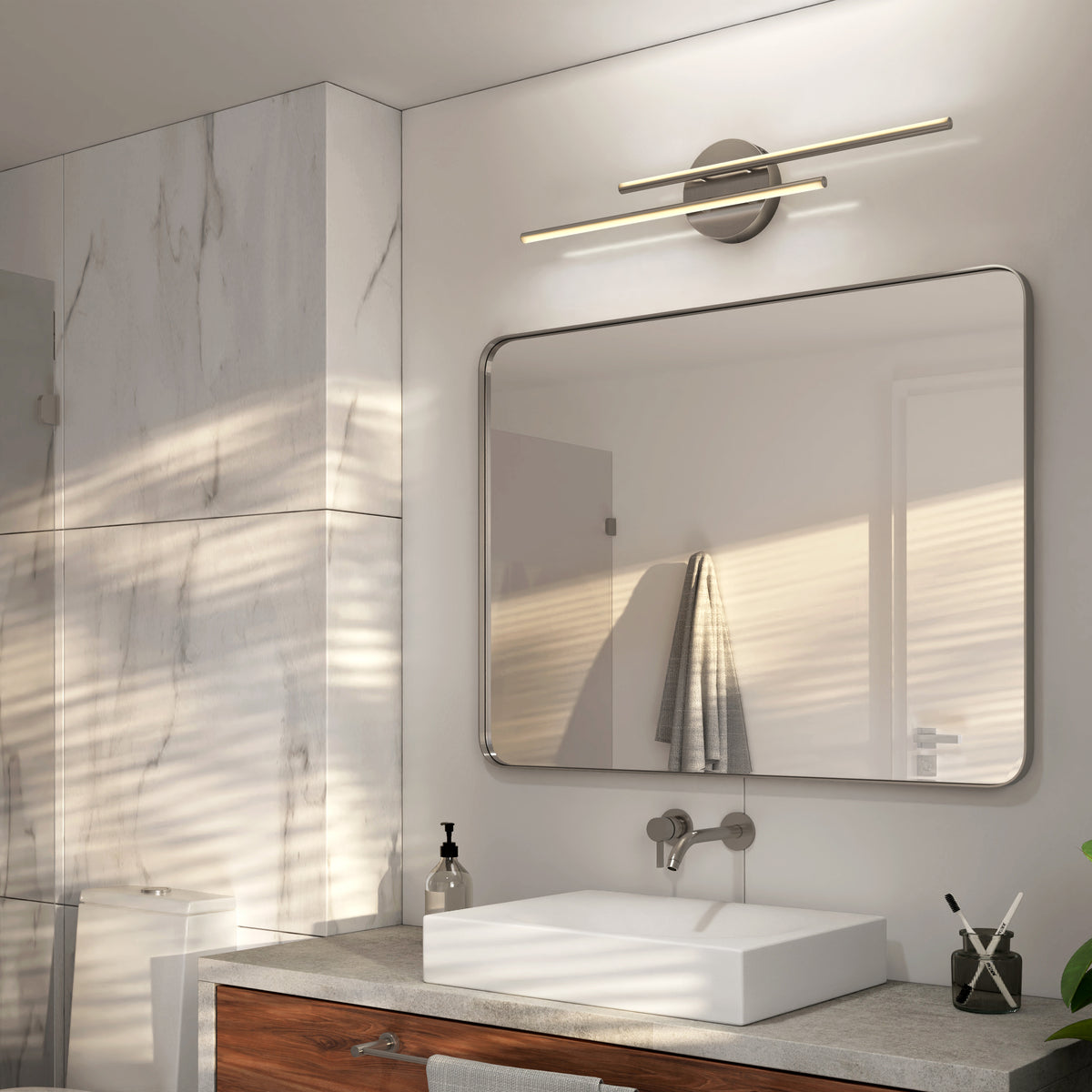 Modern bathroom with a large rectangular mirror, wall-mounted double light fixture, white sink on a wooden vanity, and marble wall tiles. Sunlight casts shadows across the space. A towel hangs near the door.