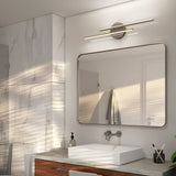 Modern bathroom with a large rectangular mirror, wall-mounted double light fixture, white sink on a wooden vanity, and marble wall tiles. Sunlight casts shadows across the space. A towel hangs near the door.