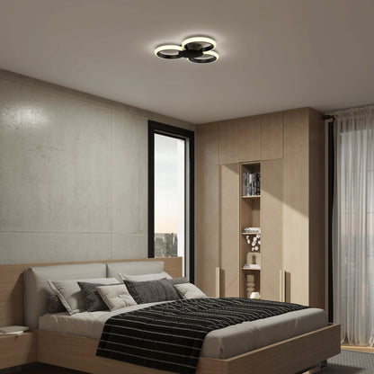 A modern bedroom features a wooden bed, black and white bedding, built-in shelving, city-view window, and the Artika Nyala 15 in. LED flush mount light 5CCT—a circular ceiling fixture. Neutral tones and minimalist decor create a cozy vibe.