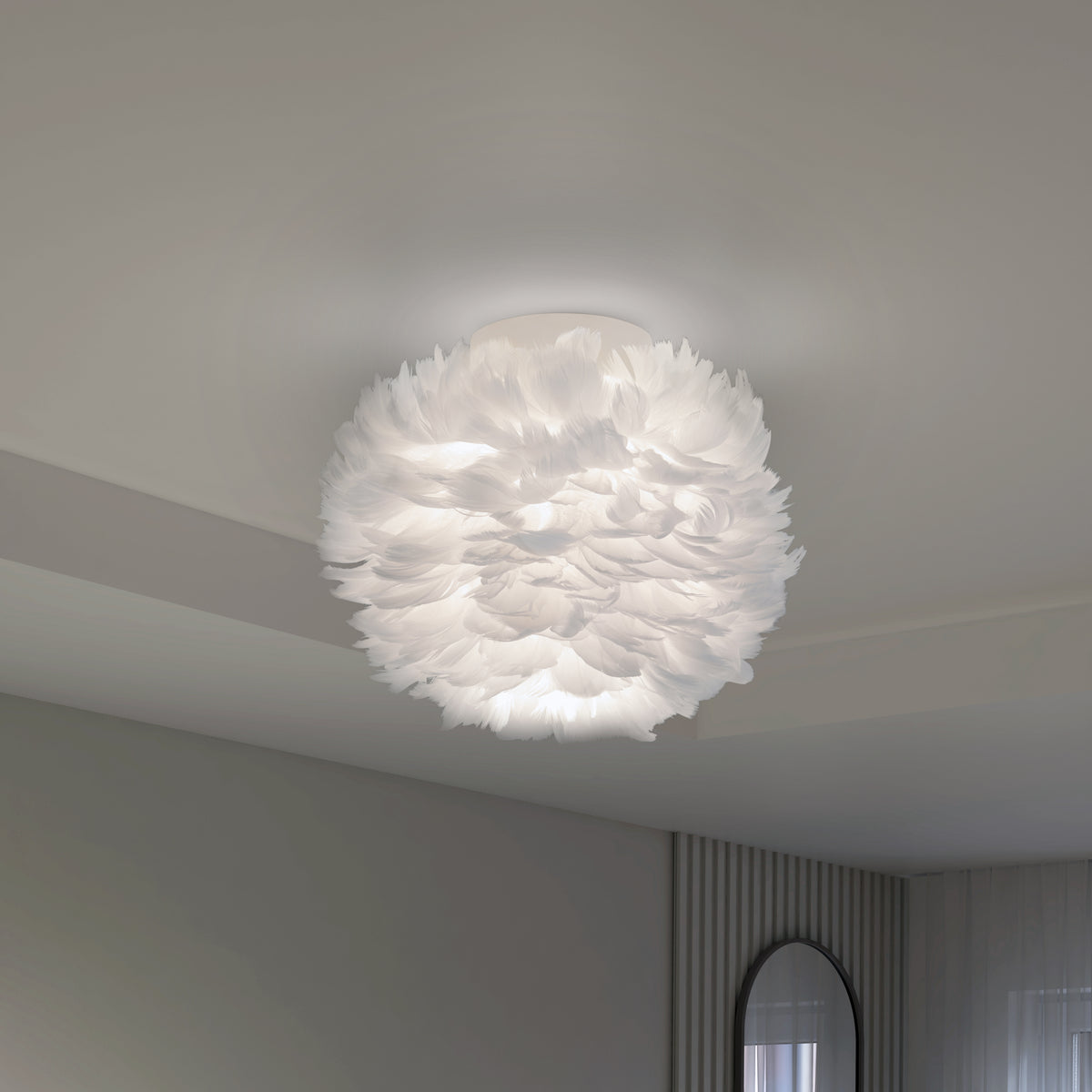 A modern ceiling light fixture with a round, fluffy white shade resembling feathers, mounted on a plain white ceiling in a minimalist room with light-colored walls and a round mirror.