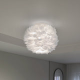 A modern ceiling light fixture with a round, fluffy white shade resembling feathers, mounted on a plain white ceiling in a minimalist room with light-colored walls and a round mirror.