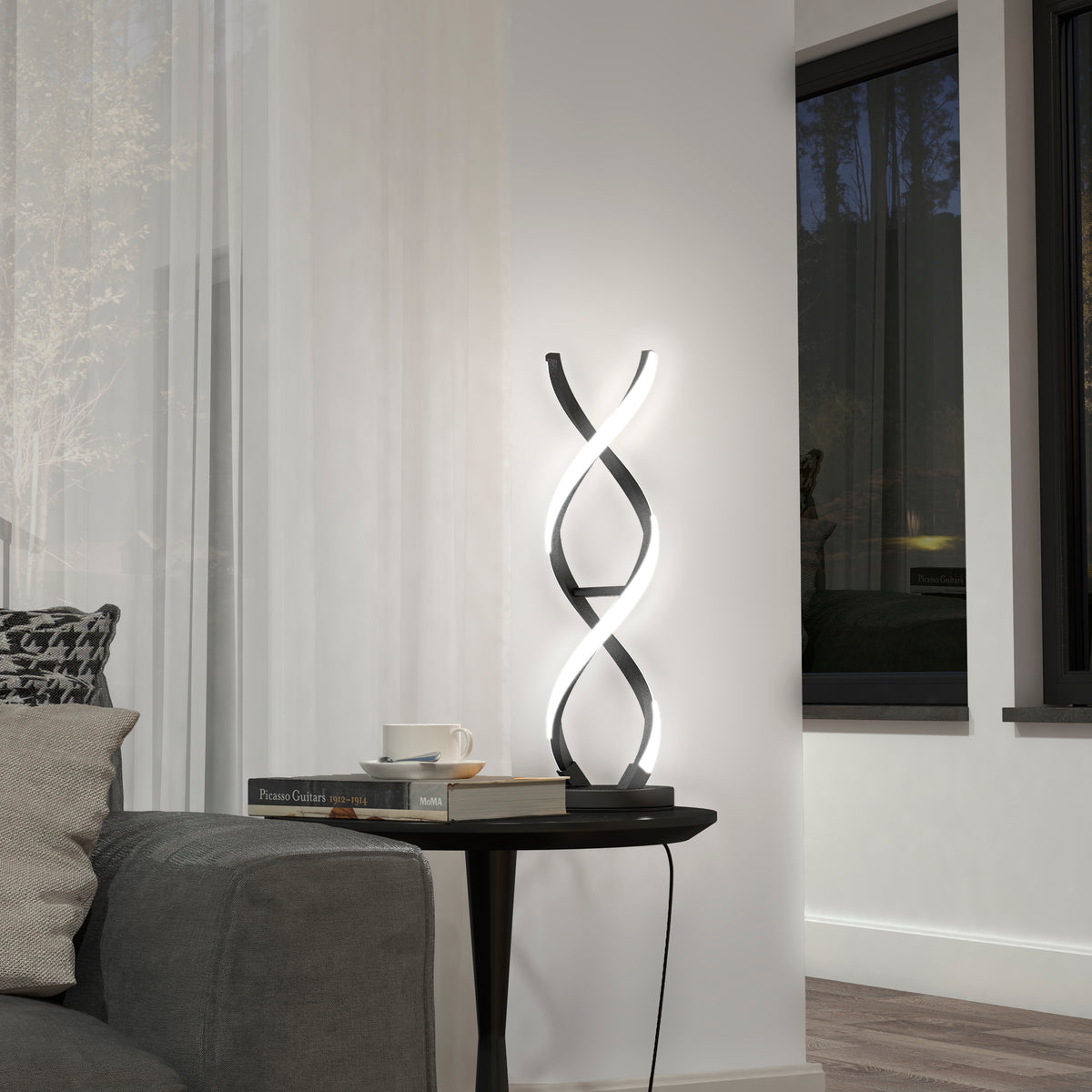 The Artika Swirl LED table lamp with a twisting design sits on a round black side table next to a gray sofa. Nearby are a cup, saucer, and books, with sheer curtains and a window softly lighting the scene.