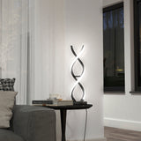 A modern spiral LED table lamp glows on a black round side table beside a gray sofa. The table holds books and a white cup. Large windows and sheer curtains are in the background.