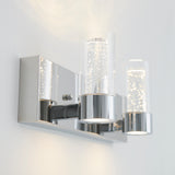 Modern chrome wall sconce with two cylindrical glass shades, each containing bubble-like details. The light is mounted on a shiny rectangular base and emits a soft, warm glow against a white wall.