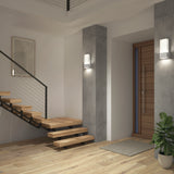 Modern entryway with a floating wooden staircase, light wood flooring, a wooden front door, concrete pillars with wall lights, a potted plant, and a framed artwork on the white wall.