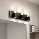 A modern bathroom light fixture with three rectangular black bases, each supporting a clear glass shade with a ribbed inner design, mounted on a light gray tiled wall above a mirror.