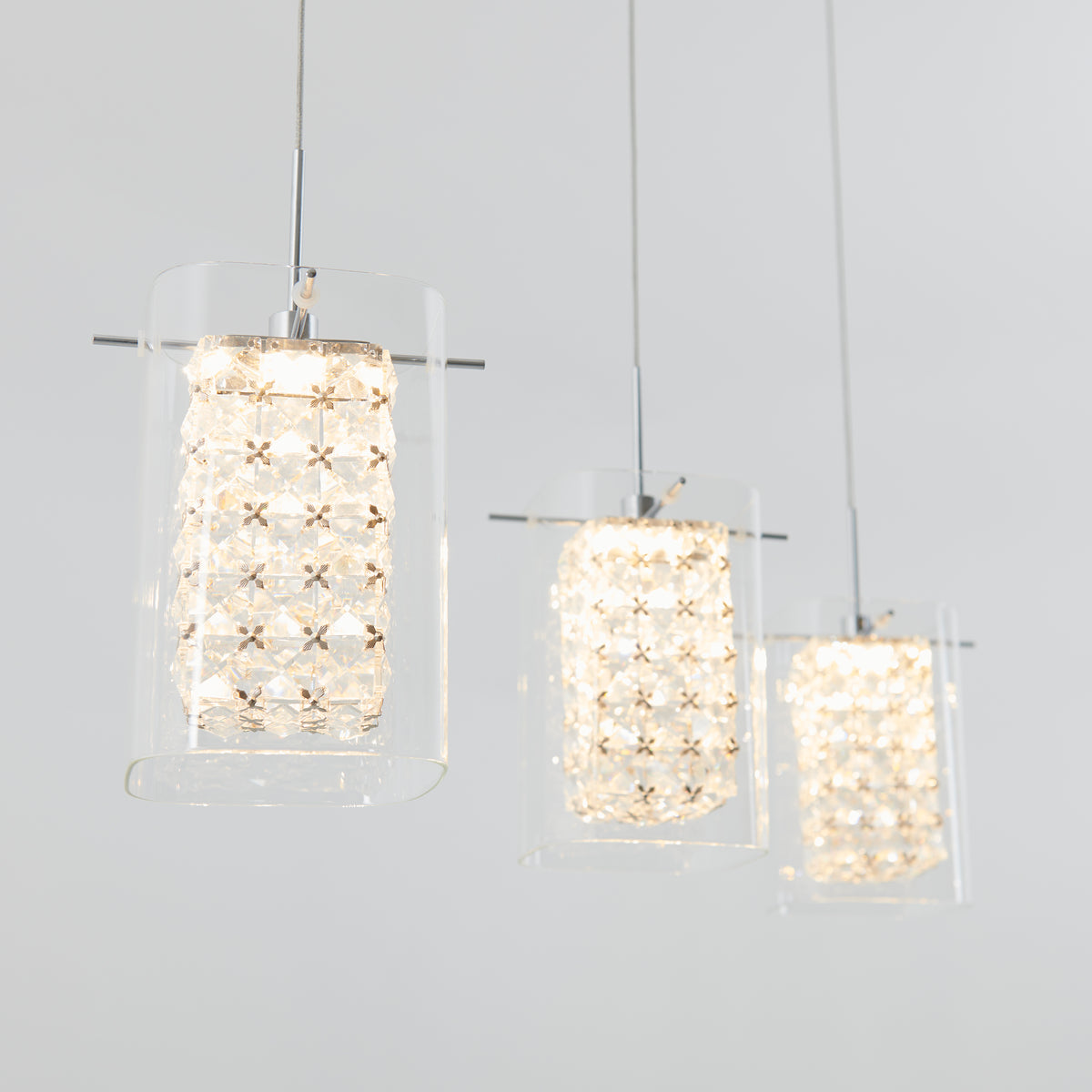 Three modern pendant lights with glass and crystal-like details hang from the ceiling against a plain light gray background, emitting a soft, warm glow.