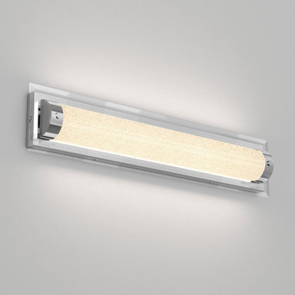 A modern wall-mounted light fixture with a cylindrical frosted glass shade and chrome end caps, emitting a soft, warm glow against a plain light gray wall.