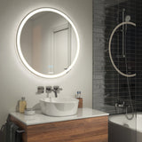 Modern bathroom with a round, backlit mirror above a white vessel sink on a wooden vanity, wall-mounted faucets, toiletries, and a bathtub with a dark tile wall and a rainfall showerhead.