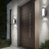 Modern wooden front door with horizontal panels, a tall metal handle, and two cylindrical wall lights on either side, mounted on a concrete exterior wall. Green foliage is visible in the foreground and background.