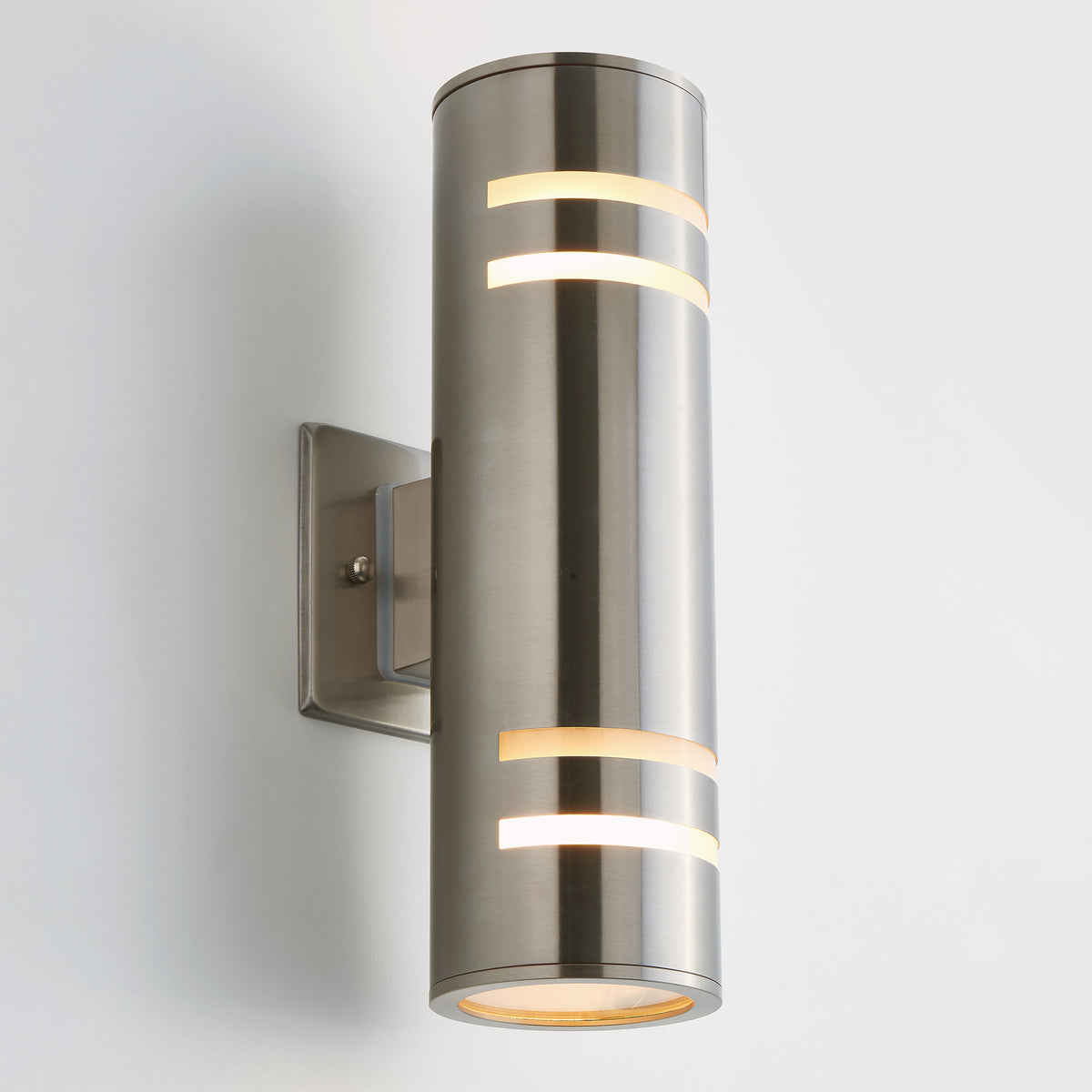 A modern, cylindrical brushed metal wall sconce with horizontal cutout slits emitting warm light, mounted on a square wall plate against a plain white background.