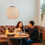 A young couple sits at a round wooden table in a cozy, modern dining nook with a tufted leather bench. They share breakfast, with juice, pastries, and fruit, under a decorative pendant light. Plants hang nearby.