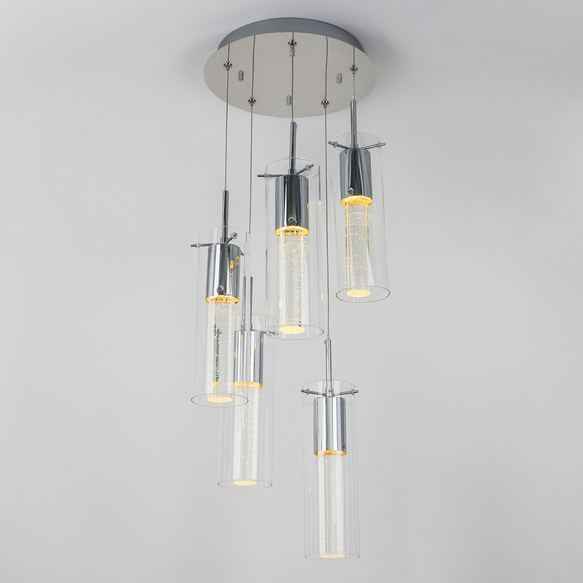 A modern ceiling light fixture with six cylindrical glass pendant lights, each containing glowing LED lights, hanging at varying lengths from a round metallic base against a white ceiling.