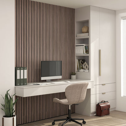 Modern home office with a white desk, computer, swivel chair, houseplant, and a Sonolok sound-absorbing wall panel kit by Artika as a vertical accent wall. Built-in shelves hold books and decor; drawers and a brown briefcase complete the scene. Sunlight streams in.