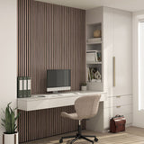 Modern home office with a floating white desk, computer, beige chair, indoor plant, and shelves with books and decor. A briefcase sits on the floor. Vertical wooden slats cover the back wall. Light streams in from the right.