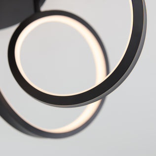 Close-up of the Artika Swirl Ribbon flush mount light, a modern fixture with two overlapping circular LED rings featuring black edges and glowing inner surfaces, set against a plain light gray background.