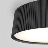 A close-up of a modern ceiling light fixture with a cylindrical, vertically-ridged black shade, glowing softly against a plain light gray background.