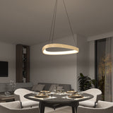 Modern dining room with a round table set for four, surrounded by light chairs. Above the table hangs an oval, wooden pendant light. In the background are a gray sofa, bookshelf, and large window with trees outside.