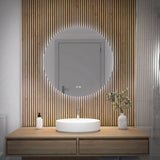 A modern bathroom with a round backlit mirror, a wooden slat accent wall, a wooden vanity with a circular white sink, a faucet, and vases with soap and green branches. A closed door is reflected in the mirror.