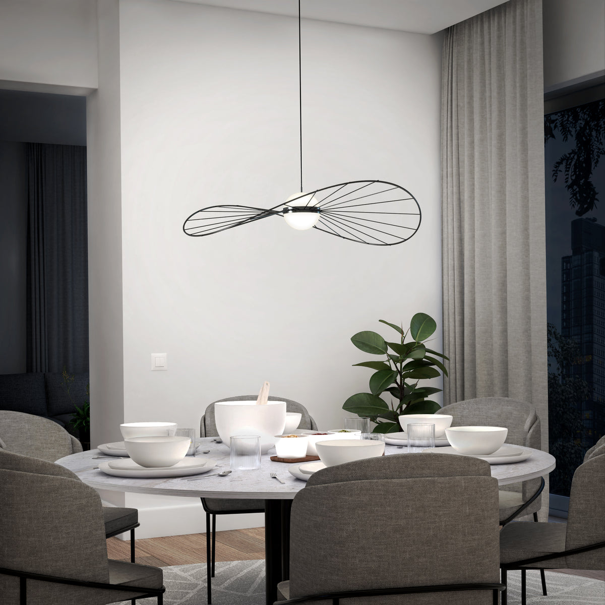 A modern dining room with a round marble table set for a meal, surrounded by gray upholstered chairs. A contemporary black wire pendant light hangs above, and a potted plant is by the window with floor-length curtains.