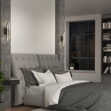 Modern bedroom with a tufted gray headboard, gray and white bedding, two wall-mounted sconce lights, a bookshelf, and a large window showing a cityscape at night. A small plant sits on the windowsill.