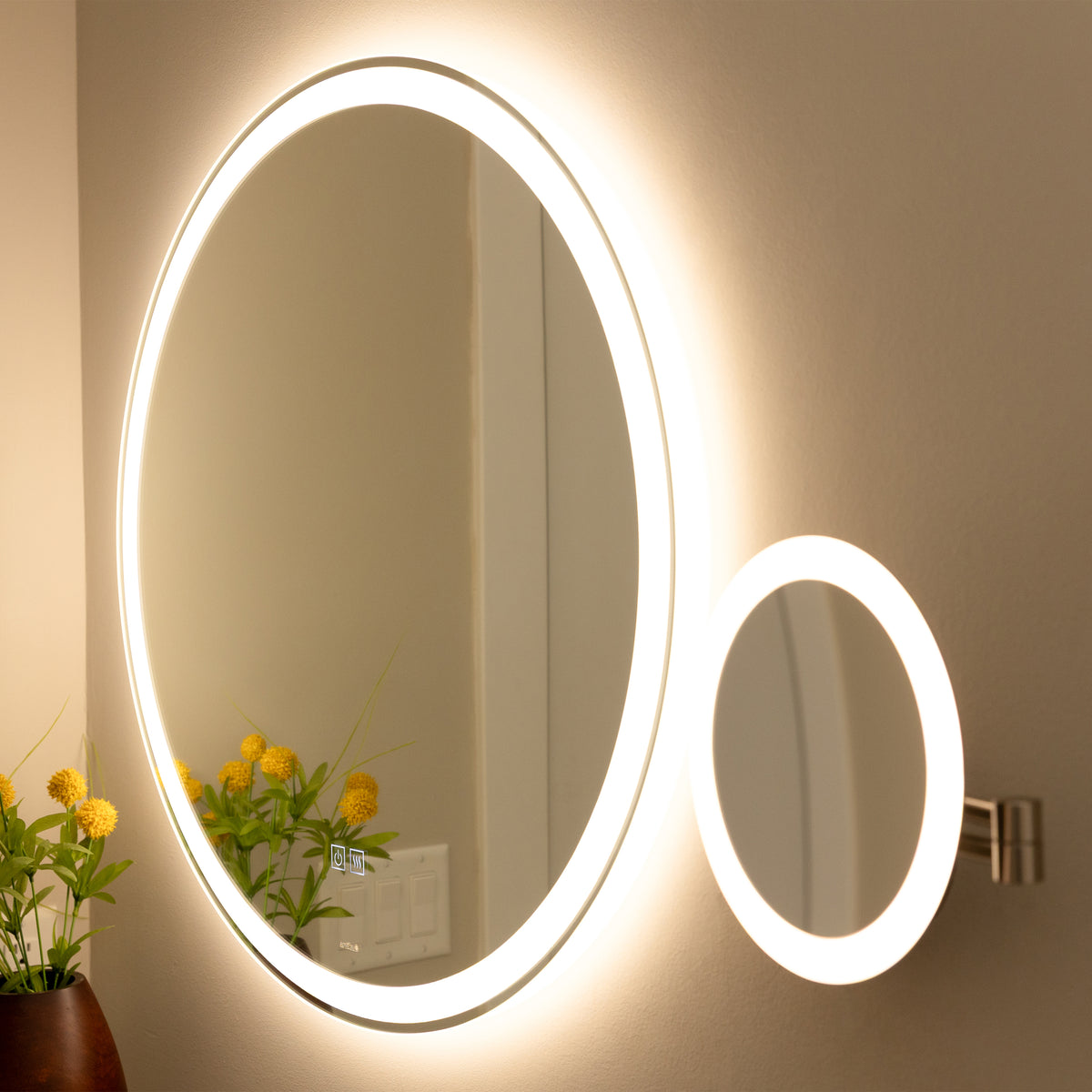 A large circular mirror with built-in LED lighting and a smaller attached magnifying mirror is mounted on a beige wall above a vase with yellow flowers. Light switches are visible in the reflection.