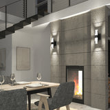 A modern dining area with a wooden table set for four, gray upholstered chairs, a concrete fireplace with a burning fire, wall sconces, and a staircase with a metal railing above.