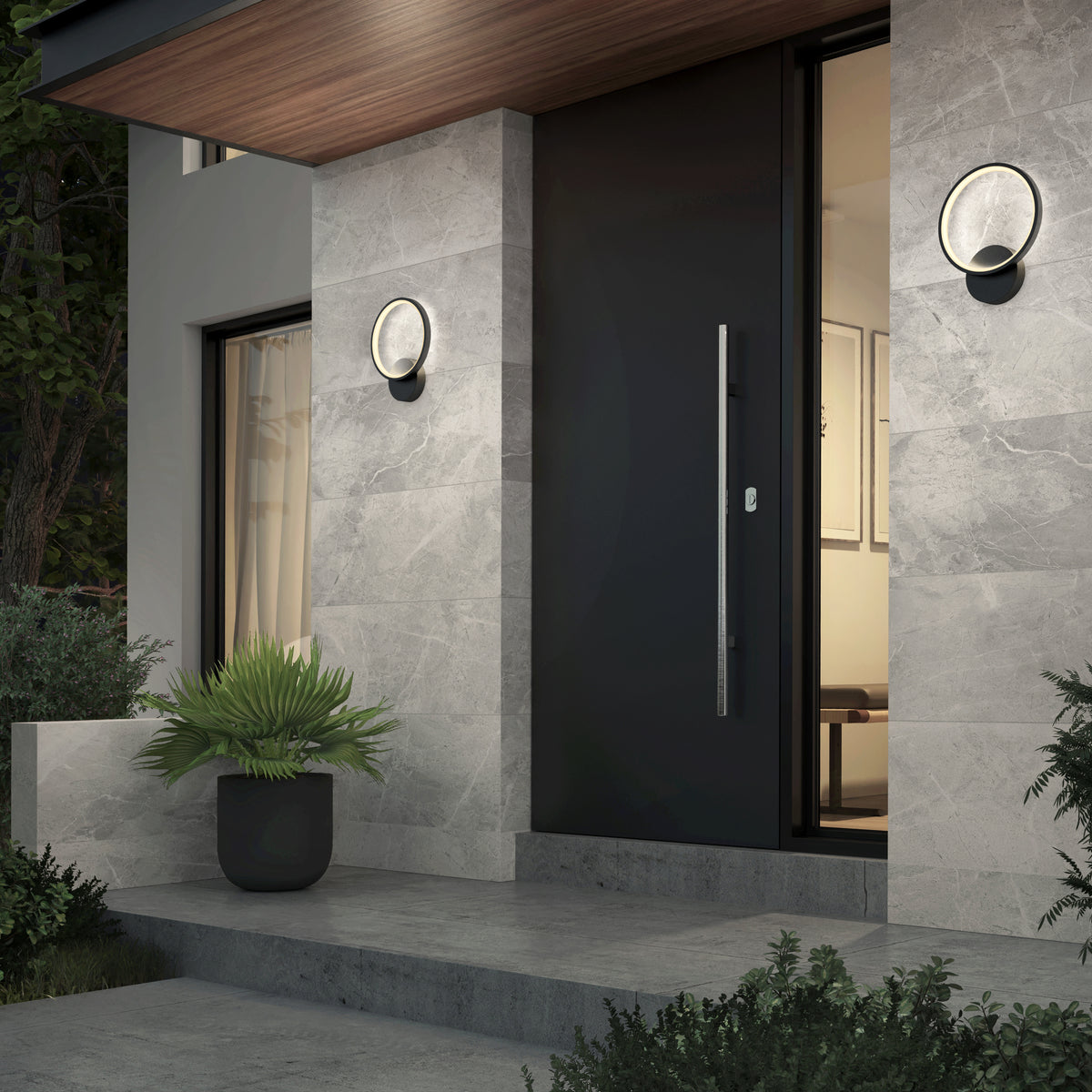 Modern house entrance with a large black door, gray stone walls, circular wall lights, a potted plant by the steps, and greenery surrounding the entryway in the evening.