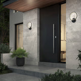 Modern house entrance with a large black door, gray stone walls, circular wall lights, a potted plant by the steps, and greenery surrounding the entryway in the evening.