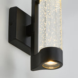 A modern black wall sconce with a clear cylindrical glass shade featuring small bubbles, emitting warm light. The fixture is mounted on a white wall.