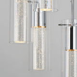 Close-up of a modern chandelier featuring cylindrical glass shades with tiny bubbles and chrome accents, each cylinder illuminated with soft, warm light. The background is a plain light gray.