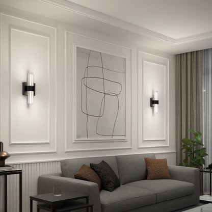 A modern living room with a gray sofa, brown cushions, minimal wall art, white paneled walls, a side table, potted plant by a large window with curtains, and Artika Dorado LED contemporary wall sconces with vertical lights.
