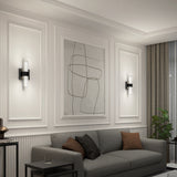A modern living room with a gray sofa, brown and gray cushions, abstract wall art, two wall-mounted light fixtures, a side table, and a potted plant near large curtains. The walls have elegant panel detailing.