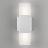 A modern wall sconce with a brushed metal rectangular center and two frosted light diffusers above and below, casting a soft glow on a light gray wall.