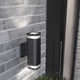 A modern black outdoor wall light fixture with two illuminated ends is mounted on a light gray brick wall next to a dark door. Green plants are visible below the fixture.