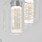 Close-up of two modern glass pendant lights with cylindrical LED bulbs inside, emitting bright white light and tiny bubbles visible in the glass, against a plain light background.