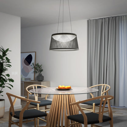A modern dining area with a glass table, four wooden chairs with black cushions, a bowl of oranges, a plant, framed art, grey curtains over the window, and an Artika Adrian LED pendant light 3 CCT illuminating the space.
