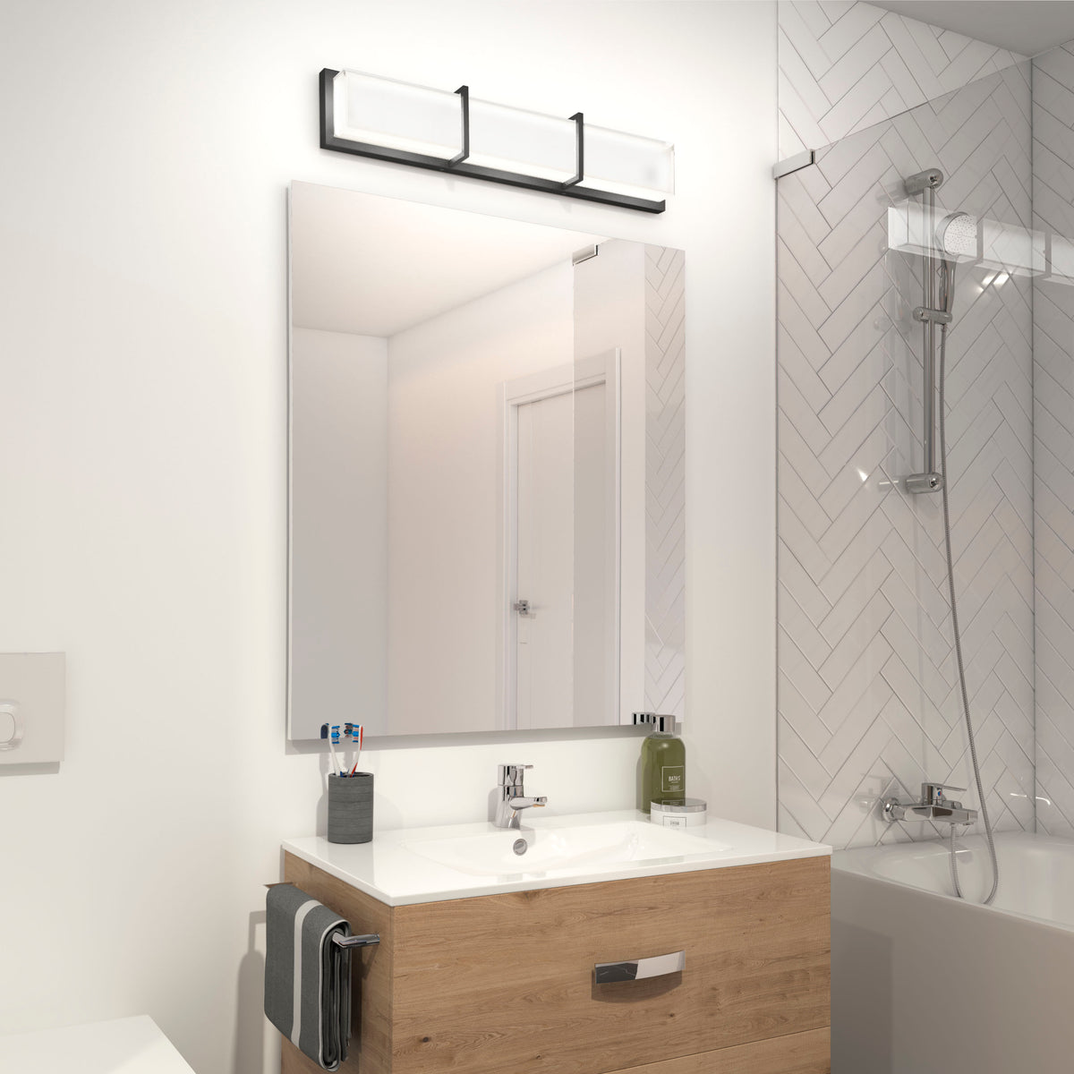 A modern bathroom with a wooden vanity, large mirror, three-light fixture above, toothbrushes in a holder, a towel hanging on the side, and a bathtub with a glass screen and herringbone tile wall.