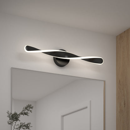 The Artika Finley LED modern bathroom vanity light 3 CCT, featuring a twisted ribbon design, is mounted above a mirror on a white wall next to a wooden panel, with a green plant in the bottom left corner.